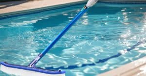 A vibrant, high-resolution photograph depicting a sparkling, crystal-clear swimming pool on a sunny day. The pool should be the central focus, showcasing its clean, inviting blue water and pristine surroundings. The photograph should have a shallow depth of field, emphasizing the clarity of the water, and should be framed horizontally for optimal banner use. The style should be realistic and photorealistic, evoking a feeling of relaxation and cleanliness. There's a blue combo pool brush attached to a telescopic pool across the pool.